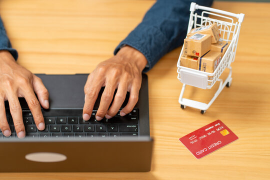 Person typing on laptop with miniature shopping cart and credit card on wooden desk