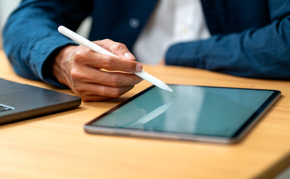 Hand using stylus on tablet with laptop beside on warm wooden desk in natural light