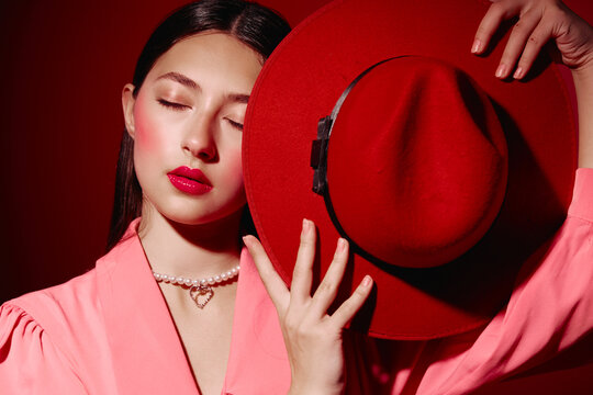 Elegant woman in a pink blazer with bold red lipstick and natural eye makeup holds a red felt hat. Soft pink and red light contrast creates a sophisticated fashion and beauty portrait.