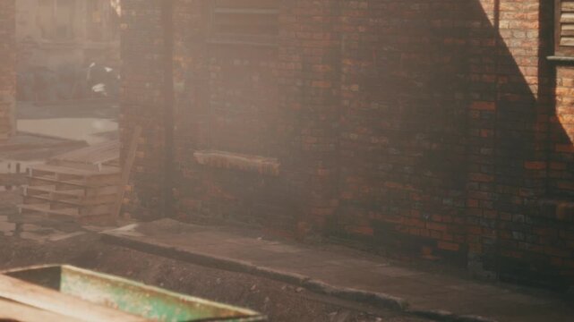 Sunlight shines on a brick wall in an urban setting. There are wooden pallets nearby and dirt on the ground. The atmosphere shows a typical work area in a city.