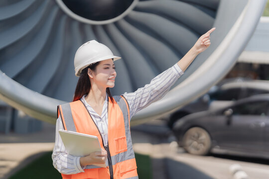 Asian female aeronautical engineer holding laptop and clipboard standing in front of airplane jet engine