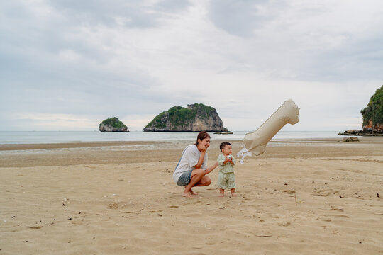 Happy Asian mother holding baby son celebrating first birthday with number 1 balloon on beach.