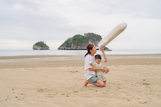 Happy Asian mother holding baby son celebrating first birthday with number 1 balloon on beach.