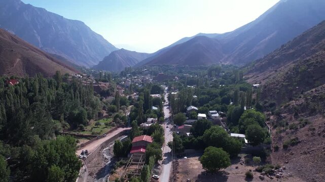 Looking down a North Road valley, a tree-lined street runs through a mountain village toward distant arid peaks. Ideal for travel, tourism, and nature stock Iran Alborz mountains valleys.