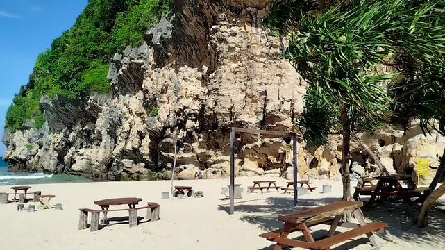 Scenic view of Lampuuk Beach in Aceh, Indonesia, featuring white sand, wooden picnic tables, turquoise ocean waves, and dramatic limestone cliffs under a blue sunny sky