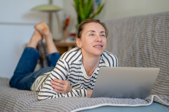 Positive thoughtful woman lying on couch browsing online on laptop relaxing at home on weekend, smiling pensive female looking away while resting on sofa with computer, leisure time and entertainment.