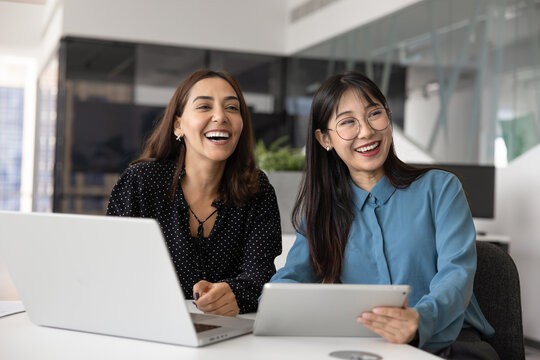 Two happy diverse women teammates, corporate employees take break seated at desk with tablet and laptop, distracted from workflow, looking away, laughing, enjoy positive, friendly talk with colleagues