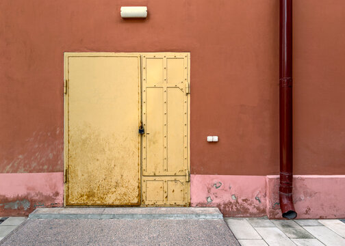 closed iron door painted in pale yellow with drainpipe on weathered red stucco wall.