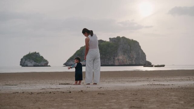 Asian mother holding hands with toddler son walking on sandy beach together.