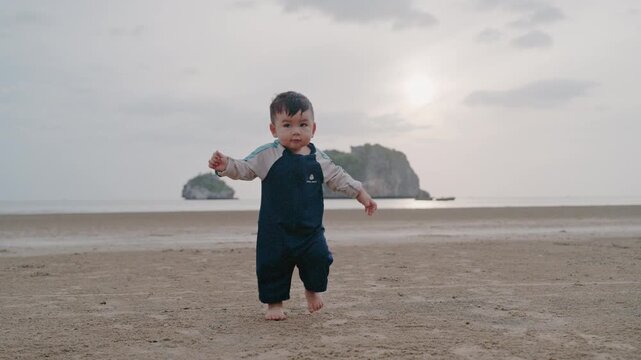 Cute Asian toddler boy taking first steps walking on sandy beach alone.
