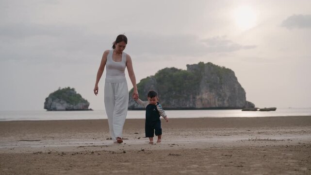 Asian mother holding hands with toddler son walking on sandy beach together.