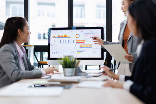 Businesswomen collaborating over financial and marketing dashboards on a monitor, reviewing charts and reports and discussing strategy, analysis and project planning in a modern office