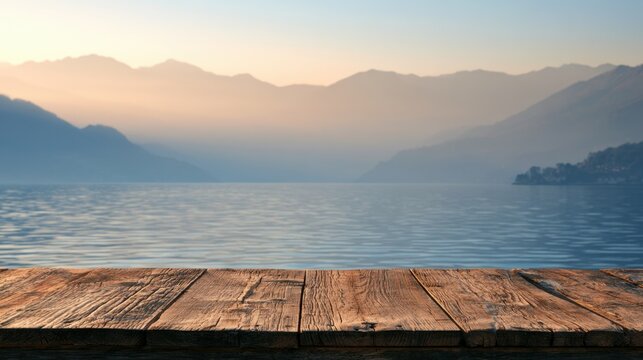 Rustic wooden table surface overlooking calm lake water with soft early morning fog and distant mountain silhouette under pastel sky light for nature and travel concept