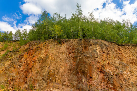 An exposed vertical rock face, an old quarry or natural cliff featuring reddish-brown, fractured rock strata topped by a green deciduous trees. Copper quarry in the Apuseni mountains - Romania
