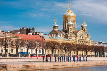 Fototapeta premium Group of people in uniform perform stretching exercises on the granite Neva embankment in Saint Petersburg, with the golden domed Assumption Church visible across the water