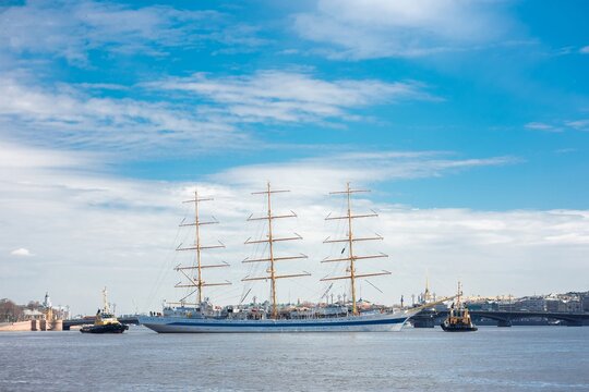 Frigate sailing on Neva River in Saint Petersburg, Russia, three-masted tall ship departing with tugboat escort under vast sky with city skyline in distance