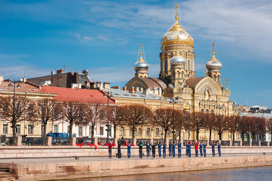 Group of people in uniform perform stretching exercises on the granite Neva embankment in Saint Petersburg, with the golden domed Assumption Church visible across the water