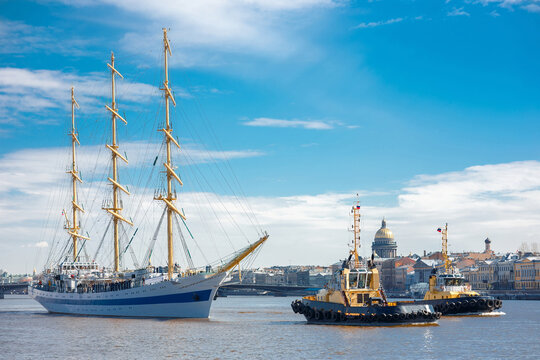 Tall sailing ship with three masts towed by tugboats on river with Saint Petersburg skyline and cathedral dome in background, maritime transportation scene