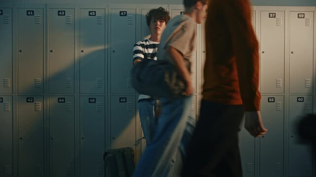 Sad Male Student Leans Against Metal Lockers in Crowded School Hallway While Classmates Walk Past. Concept of Loneliness, Social Anxiety and Emotional Struggles of Teenage Life in High School.