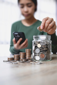Woman putting a coin into a glass jar for saving money, with stacks of coins on a wooden table. Financial planning concept.