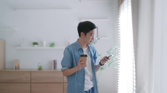 Happy Asian man drinking hot coffee in the morning using the phone while standing in the living room at home. Handsome young man walking around with a cup of hot drink, looks at a smartphone