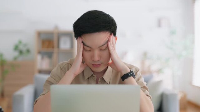 Young Asian man tired bored sad upset fatigued working on laptop in living room at home