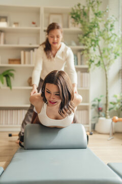 A personal trainer standing and assisting a woman's leg stretch while she performs a prone extension exercise on a Pilates arc barrel, in a bright sunlit studio.