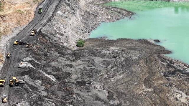 An aerial view of a former coal mine pit filled with blue water shows haul trucks operating on a road at the lake's edge, with plumes of smoke rising nearby
