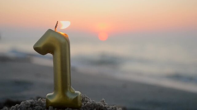 Numeral one from yellow gold color paraffin candle wax burns on sandy beach of sea shore at dawn sunset close-up. Paraffin candle burning near sea. Concept holiday Birthday New Year party celebrating