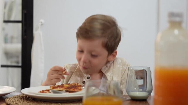 Cute little boy eating pizza at dining table at home, happy child enjoying delicious pizza for meal indoors, little boy having lunch and eating pizza with appetite