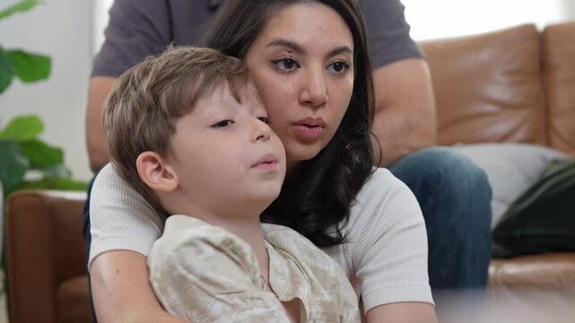Mother and son watching television and relaxing on sofa at home, caring mother hugging little boy while watching tv in living room, family watching tv program together on couch indoors