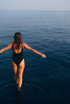 Woman in black swimsuit wading in calm turquoise sea at golden sunset