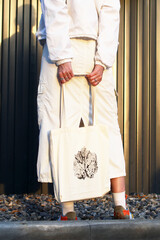 A young woman stands holding a tote bag with a leaf design. She wears a long white skirt and colorful shoes, with a minimalist background of vertical lines. © Nadezhda Zaitceva