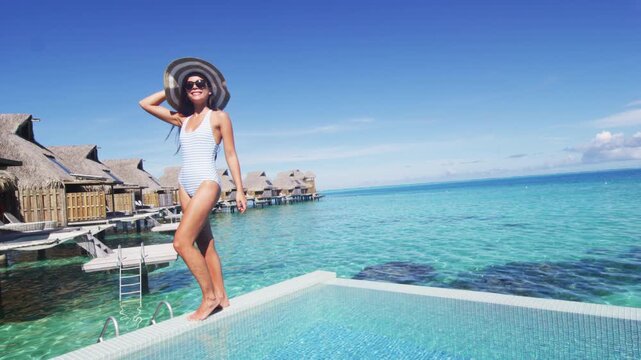 Travel vacation tourist woman relaxing at infinity pool in French Polynesia. Happy woman in swimsuit and sun hat over blue ocean at luxury resort. Raw vertical video of real woman without makeup