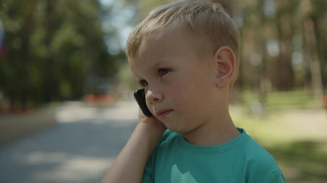 Small baby using a small phone in a park. Kid talking on an old telephone outside. Young boy holding a black phone to his ear. Child speaking by a cellular phone device in a park.