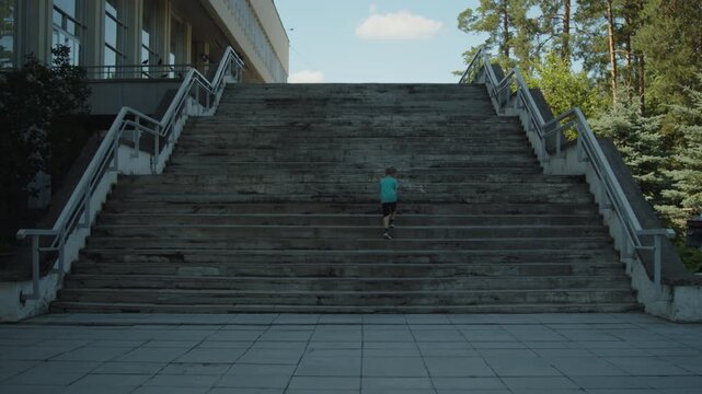 Wide shot of a young boy running up a massive concrete staircase. Active child climbing tall stone steps outside a brutalist public building on a sunny day.