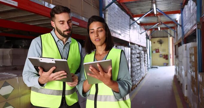 Two Latin colleagues working on inventory in warehouse together, using tablet and consignment document on clipboard, talking, discussing storage organization, getting happy, smiling