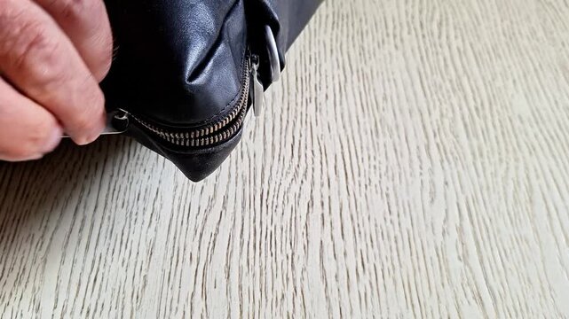 Hand opening a black leather bag zipper on a wooden floor indoors under soft natural light. Everyday lifestyle detail showing movement, texture, and casual home organization or travel prep