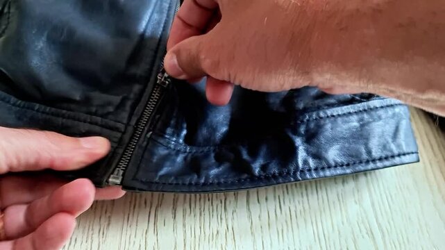 Black leather jacket zipper being fastened in a close indoor view, showing leather texture, clothing detail, and everyday fashion movement under soft natural light in a casual lifestyle scene