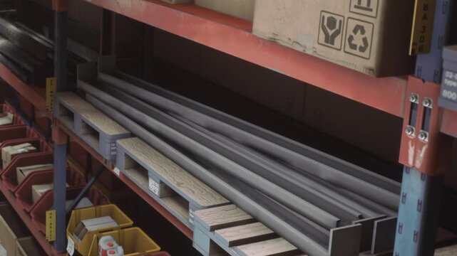Workers sort and store metal beams on shelves in a warehouse. The scene shows various boxes and containers organized alongside the metal materials.
