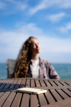 Smartphone in the foreground with a woman relaxing by the sea in soft focus behind it. Concept of digital detox, disconnecting from technology, mindfulness and finding balance in everyday life.