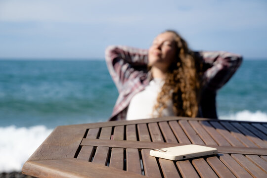 Smartphone in the foreground and a woman relaxing by the seaside in the background. Conceptual lifestyle image about digital detox, disconnecting from social media, enjoying time away from technology.