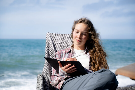 Young woman keeping a gratitude journal by the sea on a sunny day, writing in a notebook while enjoying a quiet outdoor moment. Mindfulness, self-care and intentional living concept.