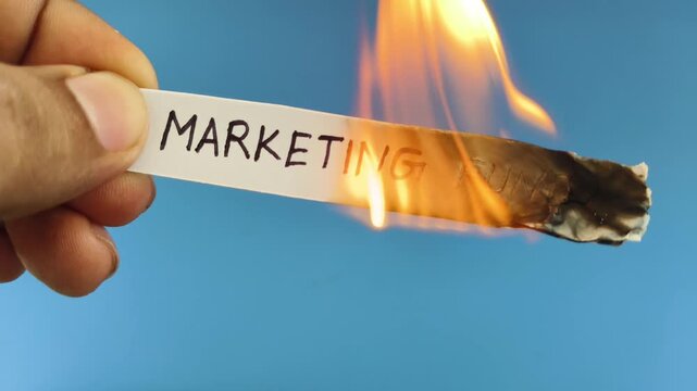 Closeup of hand holding white paper note with marketing fund word or text written on the surface with burning fire flame. Business advertising budget cost loss, waste or failure symbolic concept.