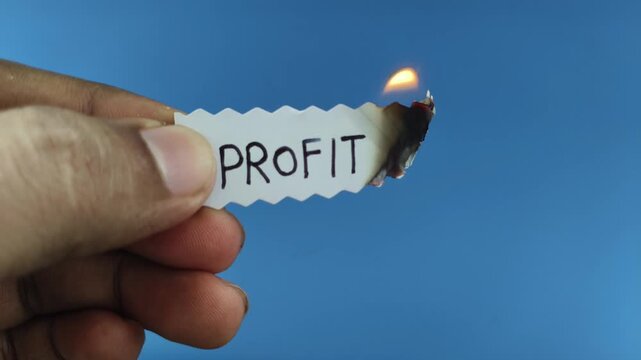 Hand hold white paper strip with profit word written on the surface with burning fire flame. Symbolizing financial loss, business risk, market pressure, economic crisis, and declining revenue concept.