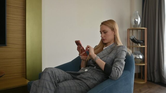 Young businesswoman relaxing in an armchair while texting on her smartphone, displaying a positive attitude and smiling during a business trip break in her modern hotel room
