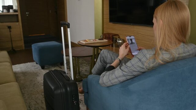 Blonde woman relaxing in her hotel room after check-in, casually scrolling through her social media feed on a smartphone while her luggage stands nearby on the carpet