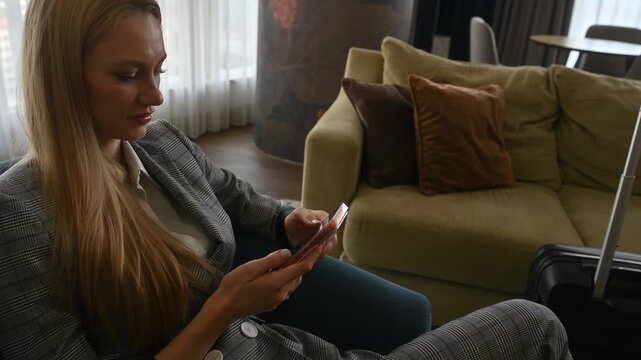 Blonde woman in a stylish suit sitting comfortably in a hotel lounge, scrolling through her smartphone while waiting with her luggage, connecting and relaxing on a business trip