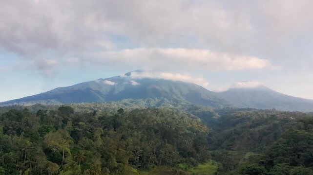 Aerial drone video of Mount Batukaru peak covered by dramatic white clouds and blue sky, with dense tropical forest in the foreground. Located in Tabanan Bali Indonesia