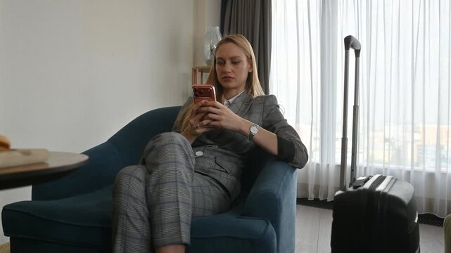 Businesswoman sitting on an armchair and texting on her mobile phone after arriving in her hotel room for a business trip, with her suitcase standing next to the window